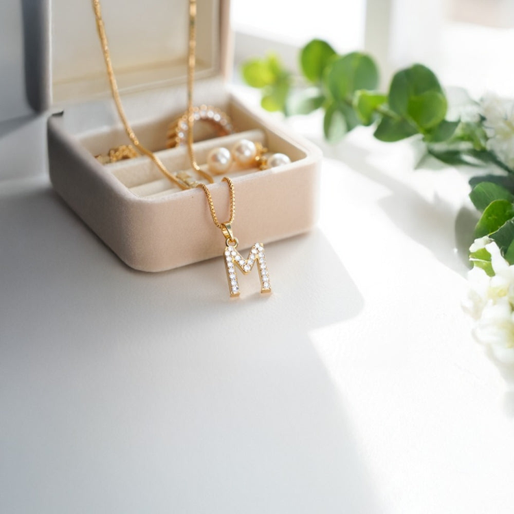 Joyero de terciopelo beige con collar enchapado en oro con dije de letra M y otras joyas organizadas.
